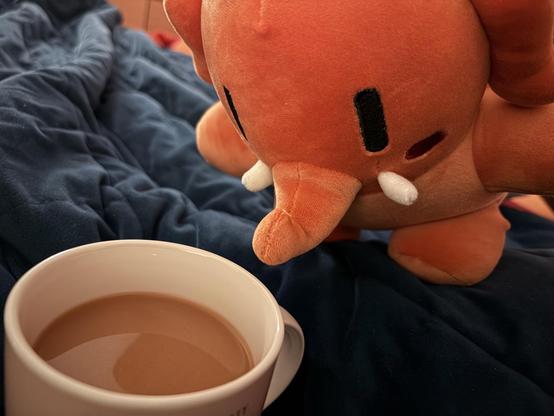 English: An orange plushtodon leans over a mug filled with coffee. The background is a blue blanket. 

Deutsch: Ein orangefarbenes Plüschtodon lehnt sich über eine Tasse, die mit Kaffee gefüllt ist. Der Hintergrund ist eine blaue Decke.