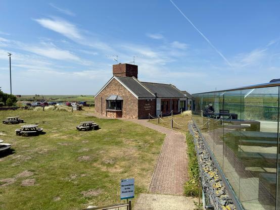 A small brick building with a central chimney and attached low extensions sits in a grassy area with several round wooden picnic tables. A narrow brick path leads to the entrance, bordered by a rope fence. To the right, a modern glass structure with stone-filled gabions reflects the surrounding landscape, where a seated person is visible. The background opens to flat marshland under a mostly clear blue sky, with a few wispy clouds and streaks of vapour trails. A line of parked cars is visible beyond the grass, alongside a tall utility pole with a single crossbar. A sign in the foreground reads “No crockery in the Picnic Area.”