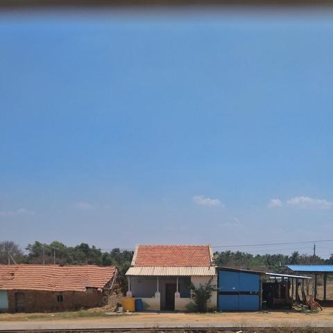 Small rural house with a red-tiled roof and a blue shed nearby, set against a clear blue sky. Sparse vegetation adds a rustic feel.