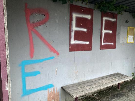Grau gestrichenes Bushaltehäuschen innen. Darauf zwei Tafeln für Plakate, die weinrot sind. Am unteren Bildrand eine Holzbank ohne Lehne. Frisch gesprüht die Buchstaben - R in Rosarot, auf die dunkelroten Tafeln zweimal E in weiß, unter dem R ein E in Türkisblau.
