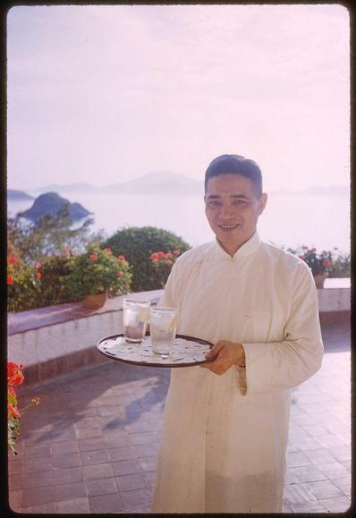 A middle-aged Asian man is smiling while standing outdoors, holding a tray with drinks on it. He's wearing traditional attire and the backdrop reveals a scenic view of water bodies and hills in Hong Kong during what appears to be daytime. The transparency shows signs of age or wear but has rich coloration indicating vibrant outdoor lighting conditions at that time.