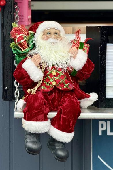 A close portrait-oriented view of a 1 foot Santa Claus figure perched on a countertop with the edge of a stack of cup lids visible next to him. He’s dressed in a dark but very vibrant red suit with wide white trim and a waist coat decorated with golden and green  glittering braiding and big gold buttons. The green sack of toys on his back is overflowing with toys, including a very ornate small red drum. He has a very full beard and big friendly eyes peering through round spectacles.
