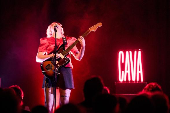 Eine Gitarristin mit roter Bluse vor rot beleuchtetem Hintergrund. Rechts von ihr eine rote Leuchtschrift mit dem Wort "CAVA". Sie hat den Kopf in den Nack geworfen, sodass ihr Gesicht nicht sichtbar ist.