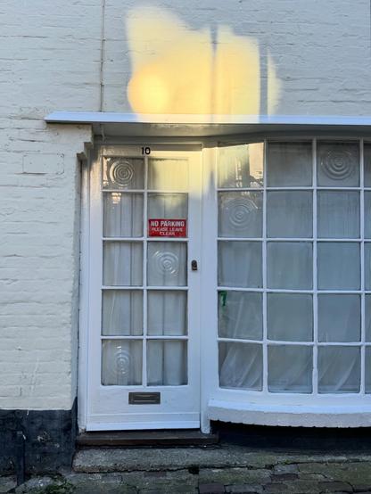 A white door that is almost entirely made up of square glass panels, next to a bay window made of the same panels. A red sign in the door says "no parking" and a brass 10 is at the top of the door. The top of the door, window, and a portion of the white brick wall around it is illuminated by a lodge of sunlight, reflecting from glass on a different building.