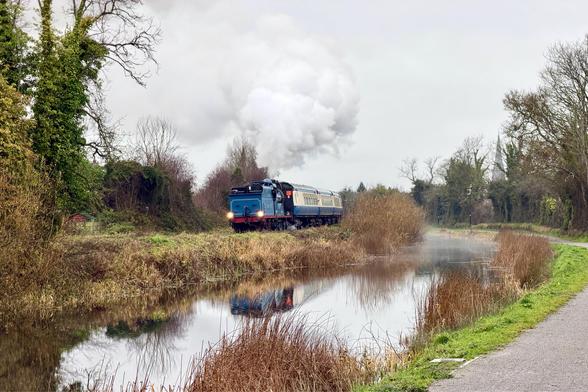 A wide landscape view of a blue steam engine running ‘backwards’ pulling a rake of cream and blue vintage carriages along the far bank of a rural canal. The big cloud of steam is clean and white and blends into a monotone grey sky above. There is a gravel path on the near-bank of the canal, and the path, canal, and railway all curve to the left as they vanish into the distance. Bare trees line the canal and the path.