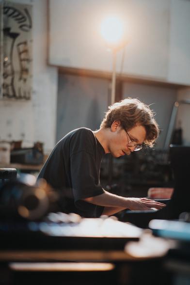 Ein Pianist im Profil spielt Klavier (das kaum zu sehen ist). Hinter dem Pianisten und über seinem Kopf befindet sich ein Scheinwerfer. Im Vordergrund sind einige unscharfe Gegenstände zu sehen. Im Hintergrund hängt links ein Gemälde, das ebenfalls unscharf ist.