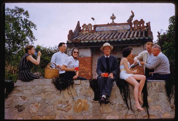 six people sitting on a brick wall in front of an asian building with tiled roof and decorative elements. two men hold red cups, another has a white cup, and one woman holds a basket next to her; the remaining three are holding teapots or serving containers. they appear engaged in conversation while enjoying tea at what seems like a picnic spot outside historical chinese architecture