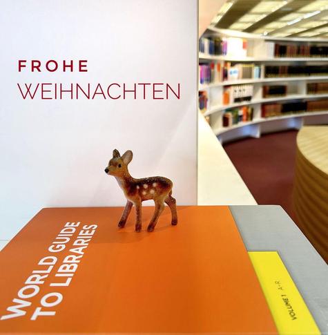 Eine kleine Figur eines Rehs steht auf einem orangefarbenen Buch mit dem Titel "World Guide to Libraries". Im Hintergrund ist eine weiße Wand mit dem Text "Frohe Weihnachten" zu sehen, und Regale voller Bücher sind sichtbar.