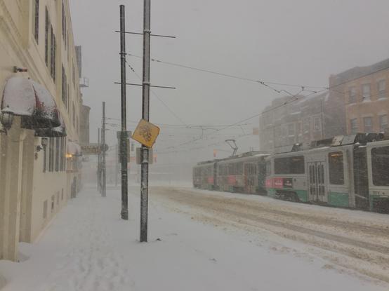 Boston’s MBTA Green Line rides by on a snowy day in the Mission Hill neighborhood. The streets are empty. Tire tracks are visible on the street and foot prints are visible on the sidewalk.