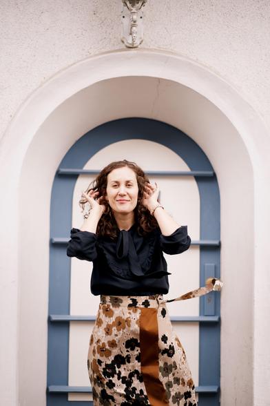 Eine offensichtlich gutgelaunte Frau steht draußen vor einer hübschen blau-weißen Holztür. Der Wind zerzaust ihr Haar, sie lächelt.