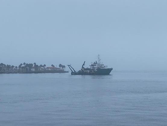 A green-hulled research vessel, the RV Weatherbird, with its bow toward the right and its stern toward the left. The A-frame, a large moveable frame attached to the stern, hangs out sub-vertically from the stern, over the flat calm, gray water. Lights shine in various places on the ship. The quarters and bridge are white, in contrast to the dark green hull which looks black in the photo. There is hardly any visible horizon due to overcast foggy conditions. There is a small peninsula to the left (aft) of the ship, with white buildings that have read tile grooves and several palm trees which are taller than the buildings.