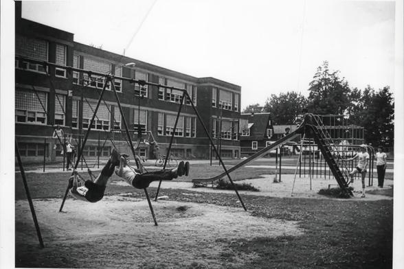 1981 black and white photograph of the Mohawk School playground in Scotia, NY. The school, two stories of brick (with a half-story windowed basement below) looms in the background, with very tall windows that are mostly glass block. In the playground there are metal-framed swings with rubber seats, children swinging on them and significant divots in the sand beneath them from years of swinging feet. A slide, monkey bars, and other climbing equipment are all visible, and there is a basketball court in the background.