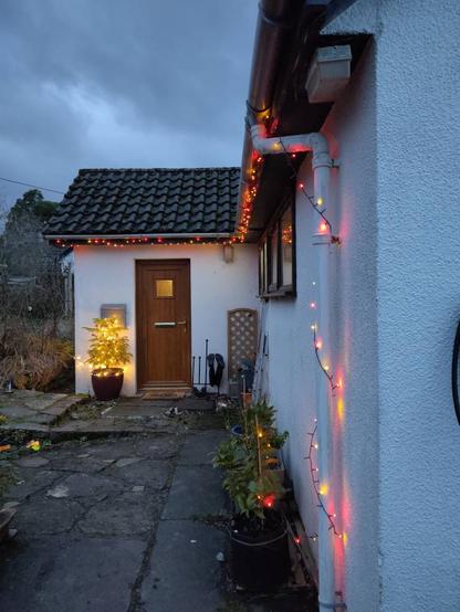 External view of the house. Colourful lights run along the roof-line and down a drain pipe.
Beside the door a small evergreen treee is adorned with white lights and various small baubles.