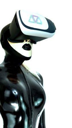 A life-sized female figure in sleek black latex stands motionless, fully encased in the glossy, skin-tight material. A VR headset covers her eyes and ears, blocking out the world, while sharply defined black lips peek beneath — slightly parted, adding a hint of eerie sensuality. The figure is posed upright, expressionless and immobilized, embodying total technological control and fetishized objectification.