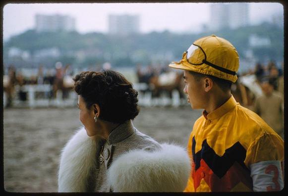 A woman in a fur coat and earrings is standing beside an Asian jockey wearing a helmet, goggles, yellow shirt with red and black stripes down the side, and number "3" on his right arm. They appear to be at horse racing track; several horses are visible behind them along with some spectators or staff members.