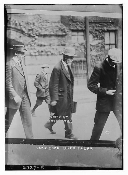 The image is a black-and-white photograph from what appears to be the early 20th century, judging by the clothing and photographic style. It shows four men walking on a street. The men are dressed in suits and hats typical of the era, with the central figure wearing a wide-brimmed hat and a long coat. The men are walking in a line, and the background features a wall with some vegetation climbing on it. The image has a caption at the bottom that reads: "PHOTO BY BOSTON TRA," and another line of text at the bottom that says: "GEO. E. COBB DRUG CLERK." The photograph is framed with a border, and there are some numbers and letters at the bottom left corner, possibly indicating a catalog or identification number for the photograph.