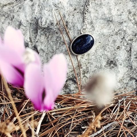 a minimal silver pendant with an oval dark blue gemstone. it has a silver chain. it's hanging on a rock and there is a pink flower on foreground