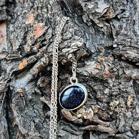 a minimal silver pendant with an oval dark blue gemstone. it has a silver chain. it's hanging on a tree