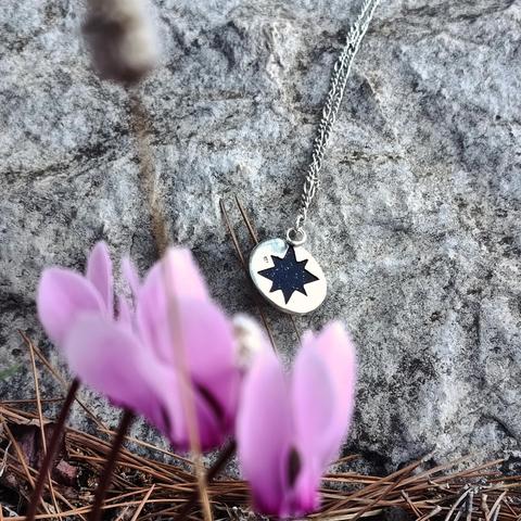 the backside of silver pendant with an oval dark blue gemstone. it has a star and and a silver chain. it's hanging on a rock and there is a pink flower on foreground