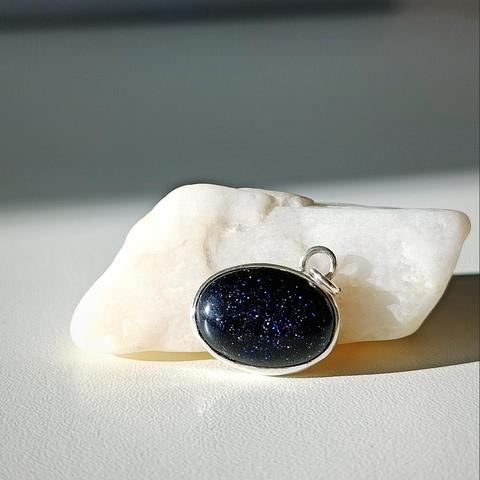 a minimal silver pendant with an oval dark blue gemstone. it is placed in front of white pebble