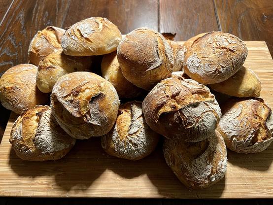 Auf einem dunklen Holztisch liegt ein Holzbrett. Darauf liegen zahlreiche frisch gebackene Brötchen mit rustikalem Ausbund.