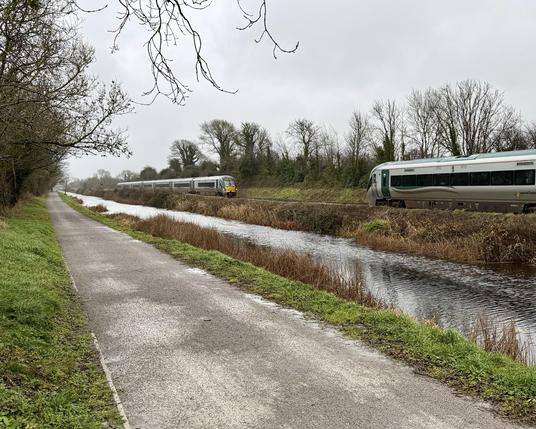 A landscape view looking up a gravel towpath in the near bank of a rural canal with a pair of railcar-type trains meeting on the far bank. The trains are both silver and dark green in colour. The scene is lined by bare trees and the sky overhead is overcast.