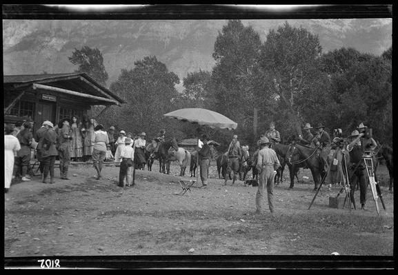 The image depicts a historic scene, likely from the late 19th century to early 20th century, involving a group of people gathered around what appears to be a movie set for a western. There are several individuals standing and seated in various poses; many wearing hats that were common during this time period. The central focus is on two horses, one with an individual mounted atop it. A large umbrella or canopy provides shade over some figures.

In the foreground, there's equipment indicative of early filmmaking techniques, such as a camera stand and possibly lights or other apparatus necessary for capturing moving images. Some individuals are wearing uniforms that resemble those from military regiments, which could suggest they're portraying soldiers in a western setting. The environment includes trees with thick foliage and what seems to be a building labeled "Post Office," further reinforcing the rural, frontier atmosphere.

The monochrome palette of this photograph adds to its vintage feel, while also emphasizing contrasts between light and shadow that were essential for early photography technology.