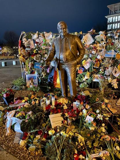 Mit vielen Blumen und anderen bunten Dingen geschmückte Stelle vor der CDU-Parteizentrale und mittendrin eine Denkmalsfigur eines Mannes im Anzug mit Schlips, die linke Hand in der Hosentasche: Walter Lübcke.