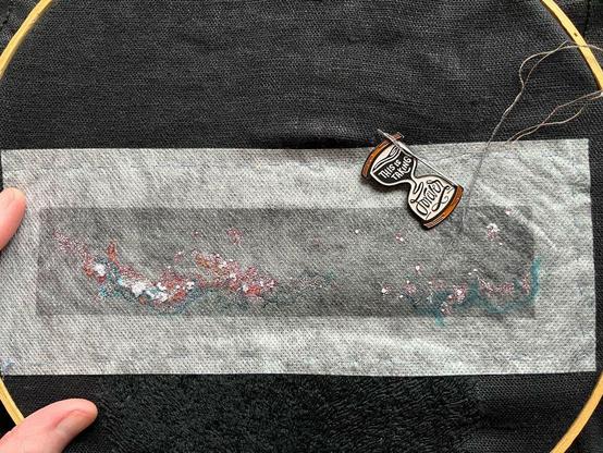 Lia’s fingers show a wooden embroidery hoop with black linen with a rectangle of dissolvable material on which is printed the Milky Way in black and white with some red and blue markings. Some tiny single strand seed stitch and French knots in white, peach, oranges, and dark browns and grays have been stitched. Below the Milky Way is black on black whipped backstitch that looks a bit like flames, it’s pubic hair. An hourglass-shaped needle minder that reads “this is taking forever” is attached to the cloth.