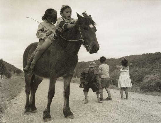 The image depicts a scene from the 1960s featuring four children outdoors in what appears to be rural New Zealand. Two young boys are riding on horseback, with one boy sitting bareback while holding onto the reins and another standing behind him for support. The woman atop the horse is wearing glasses and has long hair that extends down her back. She's also donned a skirt as she rides along.
Behind them stand three other children: two boys who are facing away from the camera, one of whom appears to be squatting or bending over while holding something in his hands; and a girl dressed in a sleeveless dress with puffed sleeves standing beside him but turned towards another child. The fourth boy is seen looking at an unseen object held by someone outside of this frame.
The setting seems pastoral, as they are on a dirt road surrounded by trees and hills, suggesting that it's either early morning or late afternoon due to the soft light quality in the photo. This photograph captures everyday life for these children from Ruatoki circa 1963, providing insight into their daily activities and environment during this period.
Additionally, there is some contextual information available regarding Maori culture: "Ruatoki" refers specifically to a tribe within New Zealand's indigenous people known as the Māori. It could imply that these children belong to or are associated with such tribes in terms of cultural identity.