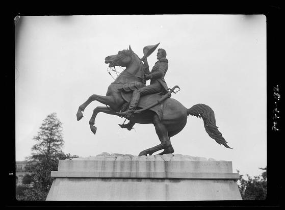 The image shows a black and white photograph of an equestrian statue, specifically titled "Clark Mills" as indicated on the pedestal. The sculpture depicts a man in mid-18th century attire atop a rearing horse. He is holding what appears to be a hat or cap with one hand while gripping reins in his other hand, which holds a sword by its scabbard. His gaze seems directed forward and slightly upward. The statue stands on a stone base that also includes the name "Clark Mills" carved into it.
The background of the photograph reveals an outdoor setting with trees visible behind the sculpture, suggesting this could be within a park or memorial area in Washington D.C., as referenced by Equestrian statues in Washington, D.C. between 1911 and 1942 from which additional information can be found regarding these sculptures.
The photographer is Arnold Genthe (1869-1942), whose work primarily consisted of photography related to art, architecture, urban life, social events, and portraiture in San Francisco. The image appears to be a nitrate negative measuring 4 x 5 inches or smaller.