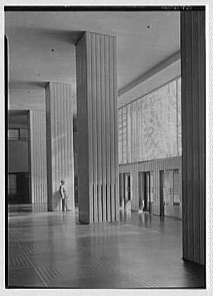 The image is a black and white photograph depicting the interior of an expansive lobby in a modernist building. The architecture features tall, narrow columns that create a rhythmic pattern throughout the space, with large windows allowing ample natural light to fill the room.
In this scene, there's only one individual visible; they appear to be standing near the center, possibly waiting or observing their surroundings. This person is wearing attire typical of mid-20th-century fashion, suggesting that the photograph was taken around 1933, as indicated in additional information provided about it being "RCA Building entrance lobby," from Rockefeller Center.
The flooring has a patterned design with lines running parallel to each other and the columns. The ceiling is not visible due to perspective; however, recessed lighting can be seen on the walls, providing illumination without any noticeable fixtures overhead.
Given that Gottschalks collection holds 1 negative: safety ;5x7 in., it indicates a professional print quality for archiving or research purposes.