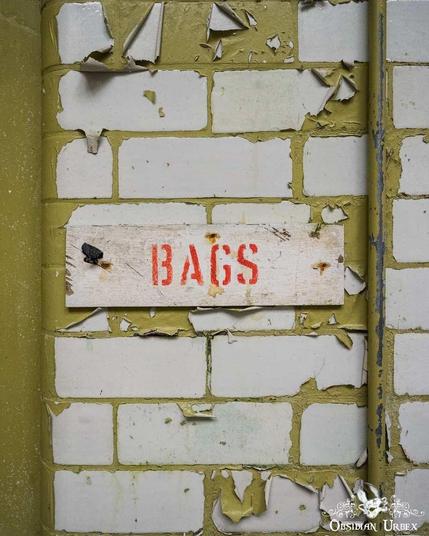An abandoned hospital laundry room sign that reads "BAGS". The peeling wall and faded color suggest age and disuse.