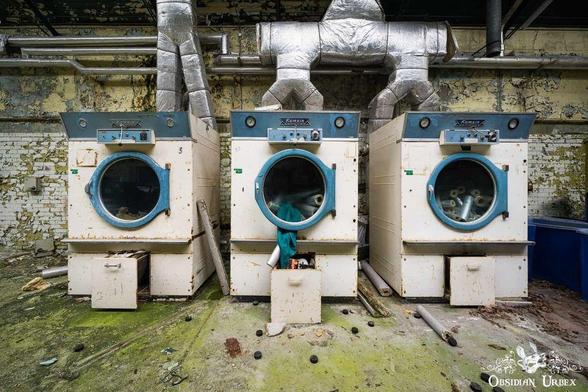 An abandoned laundry room features three decaying washers labeled "Kamsin" beneath metal ductwork. The dirty, green floor and peeling walls create a striking contrast.