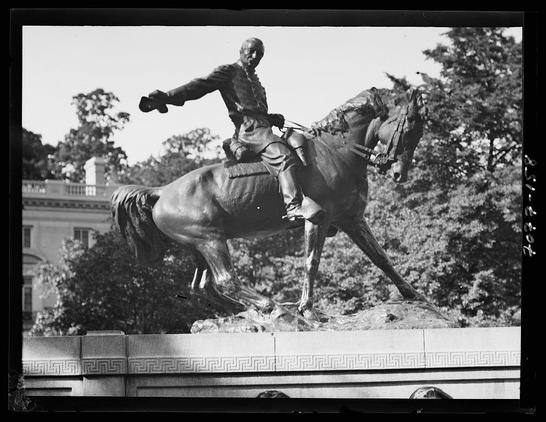 The image depicts a black and white photograph of an equestrian statue, showing a man in military attire riding on the back of a rearing horse. The figure appears to be mounted atop a stone pedestal with Greek key patterns at its base. In the background, classical architecture is visible, suggesting this statue may be located within or near such buildings. Trees and clear skies are also present in the backdrop. There's an indication that additional information about equestrian statues in Washington D.C., particularly between 1911-1942, can be found related to this photograph.