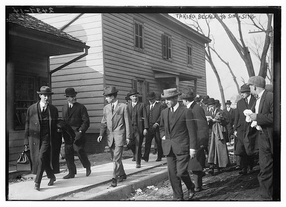 The image is a historical black-and-white photograph showing a group of individuals, likely from the early 20th century, dressed in formal attire such as suits, hats, and overcoats. They appear to be walking in a line, possibly as part of a procession or a group outing. The individuals are wearing suits with ties and some are carrying briefcases or other items. The setting is outdoors, with a wooden building in the background, which has a porch and shutters on the windows. The ground appears to be unpaved, and there are some bare trees visible, suggesting the photo was taken during a colder season. The text at the top of the image reads "TAKING BECKER TO SING-SING," which may indicate the context of the photograph, possibly related to a legal or transportation event. The overall atmosphere of the image conveys a sense of formality and purpose.