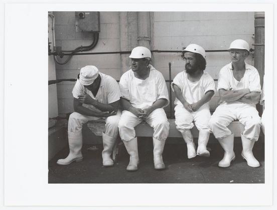 The black and white photograph features four men seated side by side on what appears to be a bench. They are dressed in similar work attire, including helmets, aprons, boots, and knee-high rubber boots, suggesting they may be employed at an industrial or food processing facility where such protective gear is required for safety reasons. The expressions of the individuals vary; one has his head bowed and seems contemplative, another looks directly towards the camera with a neutral expression, while two others have their arms crossed in what could be interpreted as defensive body language. Behind them, there's an industrial setting with visible pipes or machinery components against a concrete wall, enhancing the context of their work environment.