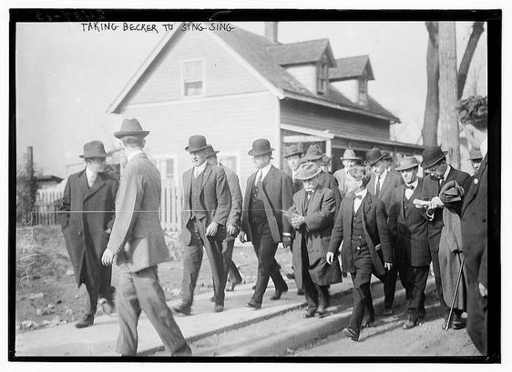 The image is a black-and-white photograph depicting a group of men walking on a sidewalk. The men are dressed in formal attire, including suits, ties, and hats, which suggests a historical context. The setting appears to be an urban or suburban area, with a two-story house in the background. The house has a pitched roof and a porch with a railing. There are trees and a fence visible, indicating a residential environment. The men are walking in a line, and the overall atmosphere of the image suggests a formal or organized event. The text at the top of the image reads "TAKING BECKER TO SING-SING," which implies that the men might be transporting someone named Becker to Sing Sing, a correctional facility. The style of the image and the clothing of the individuals suggest it is from the early 20th century.