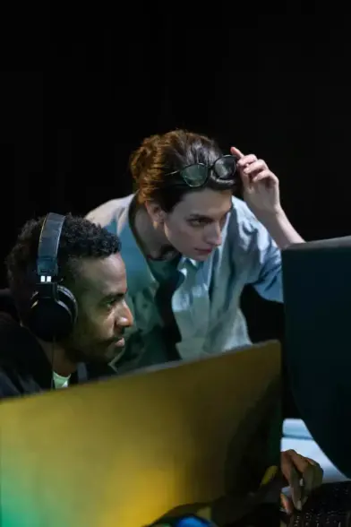 Zwei Personen konzentrieren sich auf einen Bildschirm. Die vordere Person trägt Kopfhörer, die hintere Person hat eine Brille auf dem Kopf und beugt sich vor, um besser zu sehen.