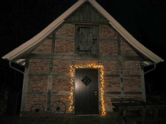 Ein kleines Fachwerkhaus mit dunklen Balken und roten Gefachen. Die kleine Holztür in der Giebelseite ist mit einer Lichterkette geschmückt. Links alte Kutschräder, rechts eine Holzbank.
