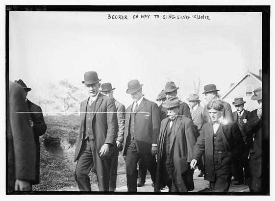 The image is a historical black-and-white photograph showing a group of men in formal attire walking together outdoors. They are dressed in suits, hats, and ties, which were typical fashion for the early 20th century. The men appear to be in a line, with their bodies oriented forward, suggesting they are walking purposefully. The setting seems to be a rural or semi-rural area, as there are trees and open land visible in the background. The sky is overcast, giving the image a somewhat somber tone. The caption at the top of the photograph reads, "BECKER ON WAY TO SING SING 10/30/12," indicating that the men are likely heading to Sing Sing, which is a correctional facility in New York State. The date "10/30/12" suggests the photograph was taken on October 30, 1912. The overall composition and attire of the individuals suggest a formal or official event.