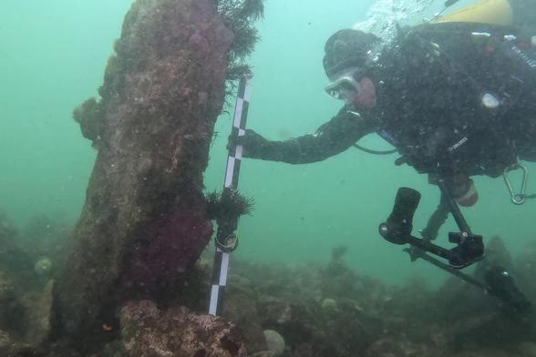 un plongeur mesure la taille d'un monolithe planté verticalement dans le fond marin.