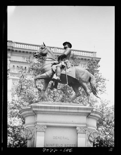 This black and white photograph captures a statue of General Winfield Scott, prominently displayed outdoors with the upper part of his monument visible. The bronze equestrian sculpture depicts Scott on horseback in mid-action, possibly representing a moment from military history or battle readiness. He is dressed in 19th-century American cavalry attire: a wide-brimmed hat, long coat, riding boots, and gloves. His right hand grasps the reins while his left holds a sword aloft. The statue stands on what appears to be a pedestal with architectural embellishments, including ornate columns at its base.

The background shows an urban environment with trees partially obscuring parts of the sculpture, suggesting that it is situated in a park or public square within a cityscape. Behind Scott's figure looms a classical-style building facade adorned with decorative elements such as balustrades and urns on pedestals atop what might be windows or cornices.

The image bears inscriptions: "GENERAL WINFIELD SCOTT" at the base of his statue, indicating identification by name; another inscription reads "7029 - 186," possibly a reference number for archival purposes. The photographer's signature is visible in the bottom left corner as well.

Notable features include the contrast between the detailed textures of Scott’s uniform and horse against the smoother surfaces of the surrounding archi [...]