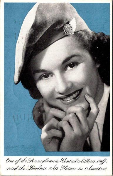This early-1940s Pennsylvania-Central Airlines postcard captures the polished, idealized image of the American “air hostess” at the dawn of commercial aviation’s first glamour age. The photograph, rendered in soft black-and-white against a bold blue background, presents a young woman in the airline’s uniform cap, complete with the PCA winged insignia—a small but proud emblem of a rapidly expanding industry. Her expression is warm and inviting, the kind of carefully cultivated friendliness airlines of the period highlighted to reassure first-time passengers and promote air travel as both modern and refined.

The caption beneath—declaring her among the “loveliest” hostesses in America—reflects the era’s highly gendered marketing strategies, when airlines routinely emphasized beauty, charm, and poise as central components of professional identity. More than just a portrait, the postcard stands as a piece of aviation advertising history: a reminder of how early airlines blended hospitality, glamour, and aspirational imagery to make flying feel both safe and sophisticated at a time when commercial aviation was still new to much of the public.