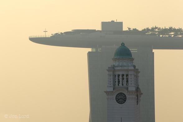 This is a colour sunrise photo in landscape format of a colonial era historic clock tower with contempory high-rise building in the background. Singapore (2015).
Below and to the right of centre is a square, white clock tower with a round dial clock in each corner, with two clock faces in view: one facing the camera and one viewed side-on. The clock face is white with the time in black Roman numerals, the clock hands also in black. The time indicated is nineteen minutes past eight. Above are square colums with a few bells visible inside. On top is a copper-green domed roof. In the background and immediatly behind, perhaps one km away, is the tall, rectangular tower of a high-rise hotel. On top of the high-rise is a large arched and slightly surfboard shaped deck that stretches from beyond the tower to the left, over the tower and off the photo to the right. The top of the deck has railings around the edge, made to look minature by the size of the deck. There are some one story structures and further to the right numerous small palm trees. Surrounding this scene and in the background is a dull orange sky, coloured by the low morning sun and probably atmospheric pollution.
The location: Marina Bay, Singapore. The clock tower belongs to the Victoria Theatre and the high-rise is the Marina Bay Sands hotel, casino and shopping centre.