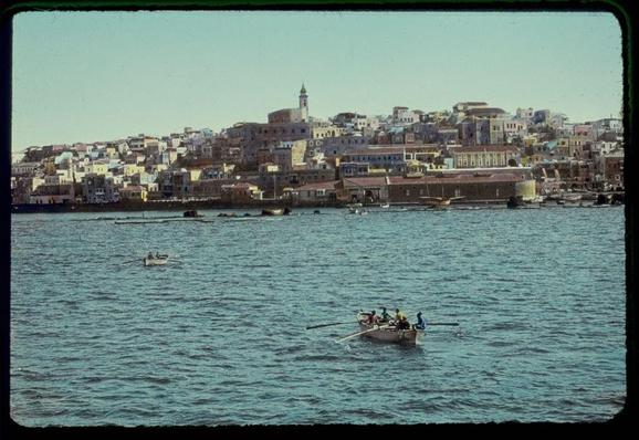The image displays a picturesque waterfront scene featuring an expansive body of water with several boats gently floating on its surface. In the background, there is a densely packed urban landscape characterized by closely clustered buildings in various shades of beige and white, some adorned with red-tiled roofs. A prominent church or mosque stands out due to its height and distinctive architectural features, including a tall tower capped with what appears to be a dome. The city extends along the shore, hinting at a bustling community nestled against the sea.
In the foreground on water are two boats: one is occupied by several individuals who seem engaged in conversation while seated alongside each other; they hold paddles and appear to row leisurely or possibly navigate through the harbor waters. Another smaller boat can be seen closer to the shoreline, with at least a couple of occupants visible. The overall atmosphere suggests a serene yet active coastal community enjoying its maritime environment amidst historical architecture.