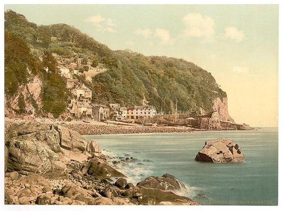 The image depicts a coastal scene with various elements in view. In the foreground, there is a rocky shoreline extending into calm blue waters where large rocks are partially submerged or protruding above water level. The middle ground shows a tranquil harbor area surrounded by buildings indicative of a small town nestled against steep cliffs adorned with dense greenery. These structures vary in size and architecture but collectively suggest an idyllic village setting, possibly from the late 19th to early 20th century judging by their style.

The background features rugged mountains or hills densely covered with trees that rise up towards a pale blue sky lightly speckled with clouds. The overall color tone of the photograph has a vintage appearance, suggesting it is an old postcard or print rather than a contemporary image. There's text on the bottom right corner which appears to be part of a caption identifying the location as Clovelly in North Devon, England.

Additional information about this particular photo can be found through online resources that mention its source from Flickr Commons and provide context regarding when it was likely taken (ca. 1890- ca. 1900). The image is part of a collection showcasing views of the British Isles specifically highlighting Clovelly, England as depicted in this postcard-like photograph by Loener.
