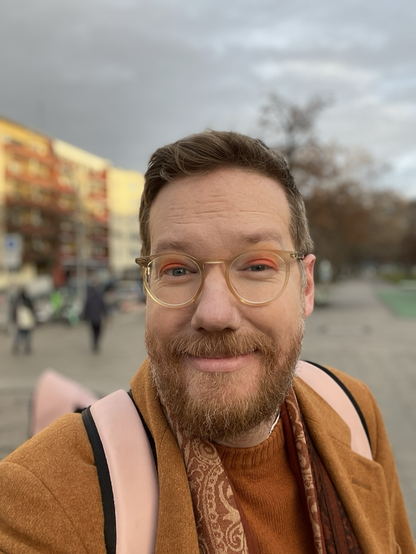 A selfie of Redfern in the street, wearing bright orange eyeshadow, an orange coat, brownish scarf, and orange sweater. They are smiling. What mischief plays on their mind today?