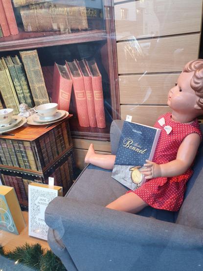 Vor einer Bücherregalatrappe sitzt eine historische Puppe mit dem Buch "Mister Bennet".