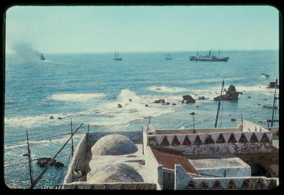 The image depicts a coastal scene with the sea taking up most of the background, showing several ships at varying distances from each other. In the foreground and middle ground, there are structures that appear to be part of an old town or cityscape near the seaside. These include domed buildings possibly indicating historical architecture, which could suggest Islamic influences given their shape resembling those found in places like Jaffa in Israel.

The sky is clear with a hint of haze over one of the ships on the horizon, and there's visible activity at sea as indicated by splashes or waves near some boats. The color palette has an aged quality to it, which could be due to fading effects from age but also adds character to the photograph giving hints that this might not have been taken in recent times.

The photo seems to capture a moment of daily life and commerce along what appears to be Jaffa's coastline, as mentioned in its caption. This scene is rich with historical connotations, suggesting it could serve as an important cultural or educational reference point for understanding the maritime activities and urban landscape around this specific location during mid-20th-century period.

The photograph has a size specification of 2 x 2 inches, implying that while it may have been taken on film given its age indicated by color saturation and clarity, such photographs we [...]